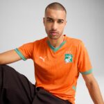 Portrait of a man wearing an orange football jersey with a Côte d'Ivoire emblem, against a light gray background.