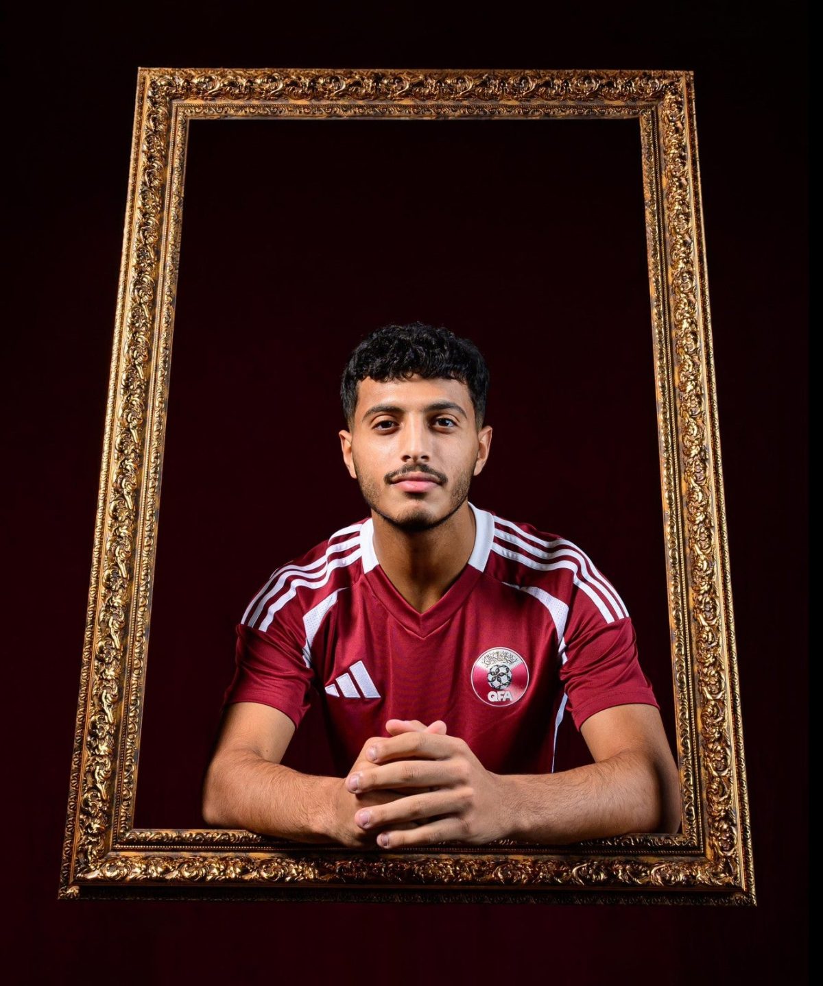 A young man in a maroon sports jersey sits with clasped hands inside a large ornate gold frame on a dark background.