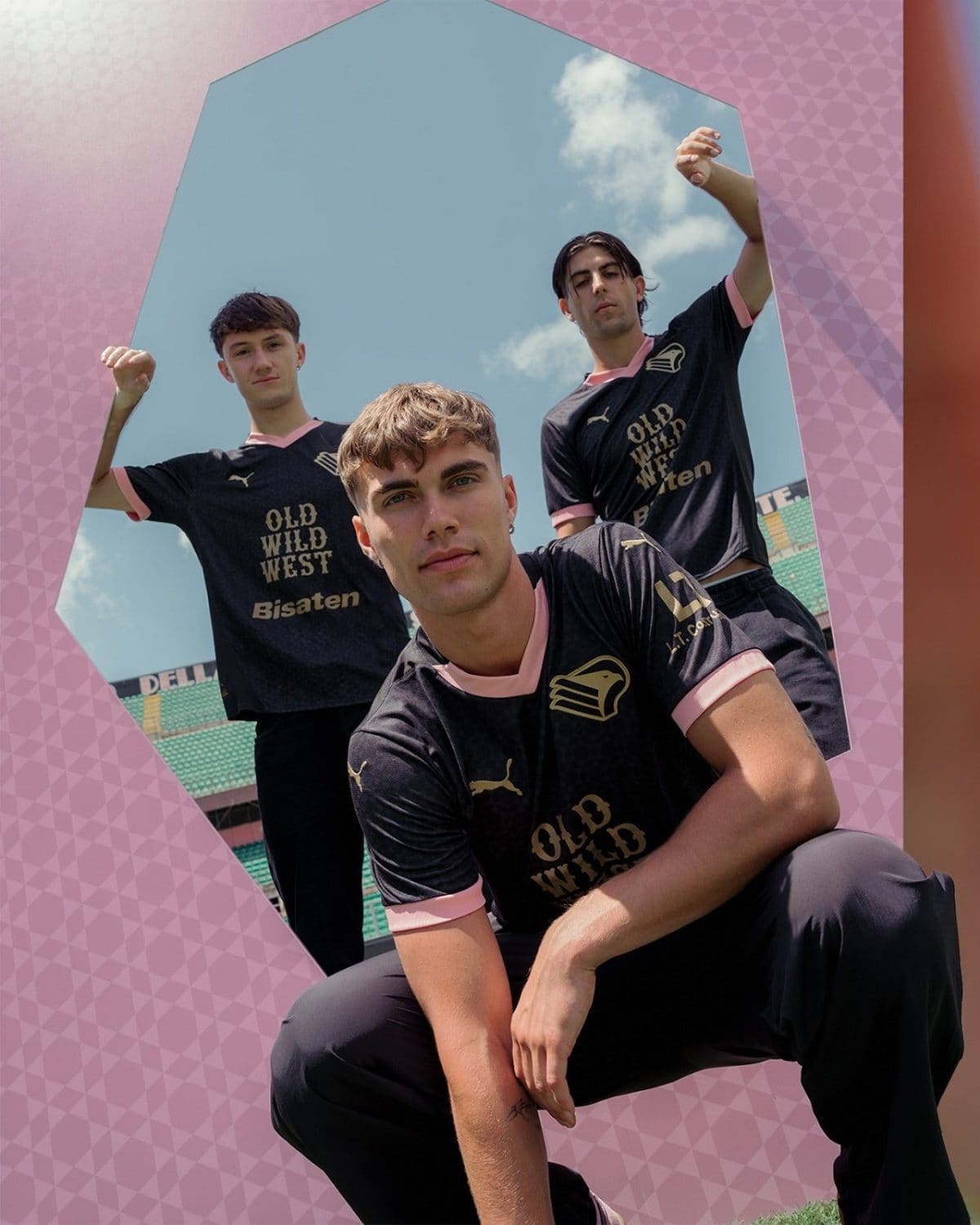 Three young men in black pink-trimmed jerseys pose together in a pink geometric frame outdoors on a sunny day at a sports field.