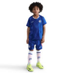 Young child wearing a Chelsea FC blue soccer kit, white socks, and neon yellow cleats, facing the camera.