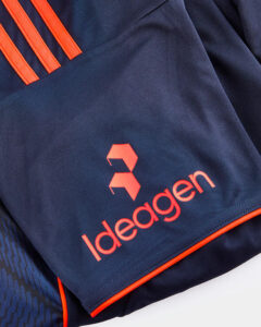 Close-up of navy athletic fabric featuring the orange Ideagen logo and three orange stripes along the shoulder/edge of the sleeve edge.