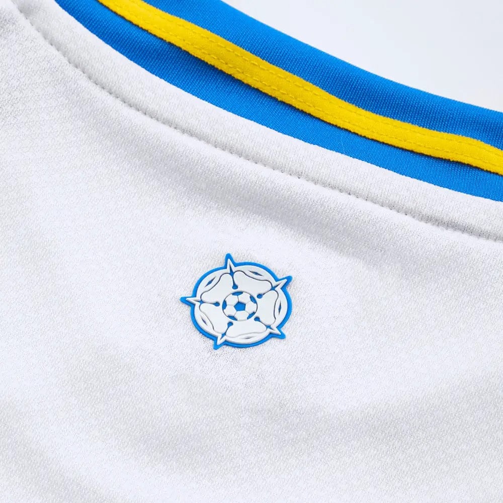 MfpUwhQKJJmdOS0 | UAE SPORT STORE Close-up of white sports jersey fabric with blue and yellow piping and a small circular pin featuring a soccer ball in blue and white.