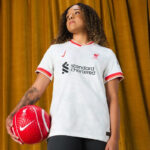 Young woman in a white Liverpool FC soccer jersey holding a red ball with a white Nike swoosh, against a gold curtain backdrop.
