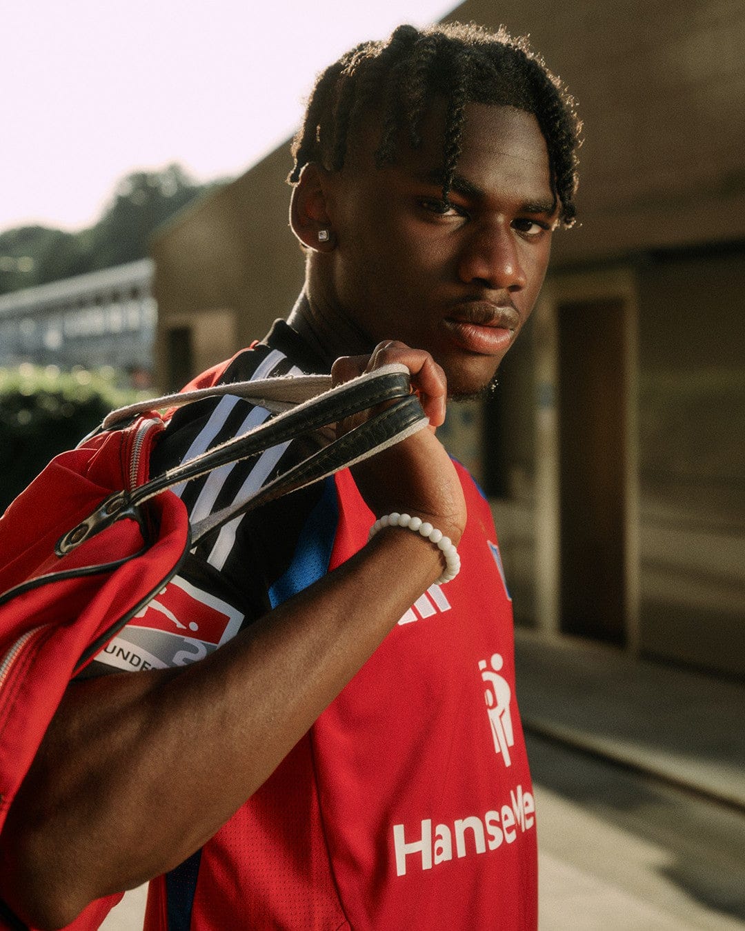 N42uSJ7Tlv3duJe | UAE SPORT STORE Young man in a red sports jersey carries a gym bag over his shoulder outdoors, looking at the camera.