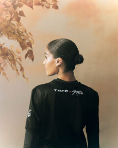 Woman with dark hair in a neat bun, seen from behind, wearing a black sports shirt with THFC x logo; autumn leaves in soft background.