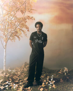 Man in black athletic outfit stands with hands behind back among autumn leaves and a pale orange sky behind him.