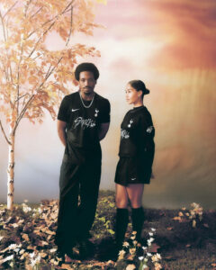 Two people stand side by side outdoors in black athletic wear with white logos amid autumn leaves and a warm sunset backdrop.