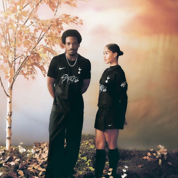Two people stand side by side outdoors in black athletic wear with white logos amid autumn leaves and a warm sunset backdrop.