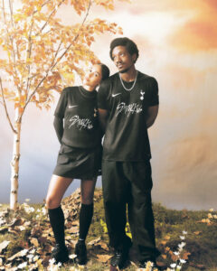 Two people in matching black athletic outfits pose together outdoors, leaning against each other among autumn leaves and a warm-toned backdrop.