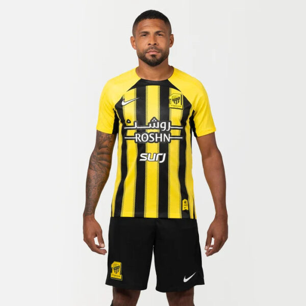 Male model wearing a yellow and black striped football jersey with ROSHN sponsor and Arabic text, plus Nike logo, paired with black shorts.