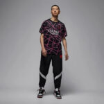 Male model wearing a black jersey with pink geometric lines and the Qatar Airways logo, paired with black joggers with reflective panels and high-top sneakers in a studio setting.