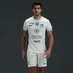 Male soccer player wearing a white jersey with blue trim and multiple sponsor logos, standing against a dark background.