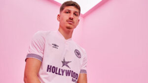 Portrait of a man in a white football jersey with Hollywood Bets logo, in a pink-lit room.
