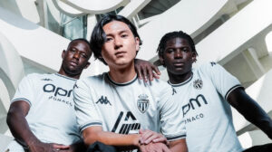Three football players pose together in light blue AS Monaco jerseys in a futuristic architectural setting.