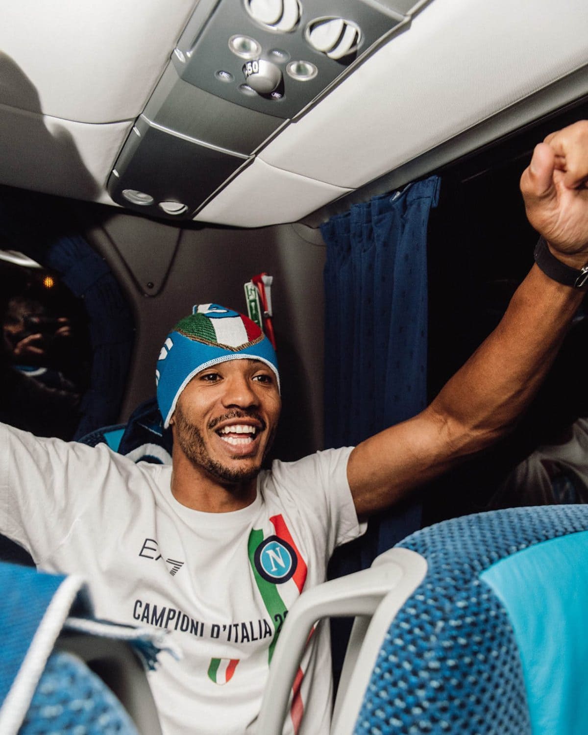 Smiling man in a white shirt with an Italy flag graphic raises his fist in celebration inside a bus with blue seats and curtains, likely a champion moment.