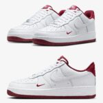 Pair of white Nike Air Force 1 sneakers with red accents on the heel and outsole set against a neutral background.