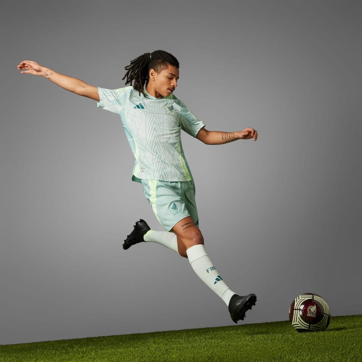Soccer player in mint green kit kicking a ball on a grassy field against a gray studio backdrop, mid‑stride.