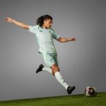 Nlbv5XBrXHSNOT0 | UAE SPORT STORE Soccer player in mint green kit kicking a ball on a grassy field against a gray studio backdrop, mid‑stride.