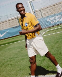 Smiling man in a yellow striped soccer jersey on a sunny field, near a goal net.