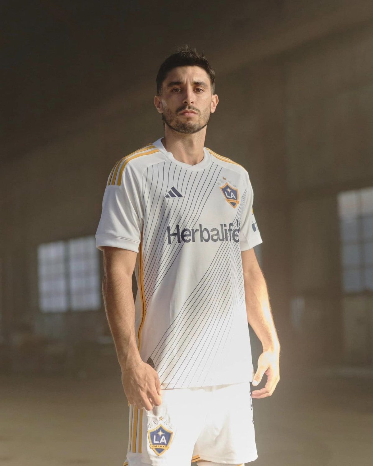 Male soccer player in a white LA Galaxy jersey with gold stripes, standing indoors and looking at the camera, braces of a dim background.