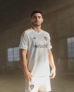 NqoNUZjmJYa6XC6 | UAE SPORT STORE Male soccer player in a white LA Galaxy jersey with gold stripes, standing indoors and looking at the camera, braces of a dim background.