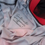 Close-up of soft light-gray fabric with a handwritten cursive message, with pink fabric and a red-trim garment nearby.