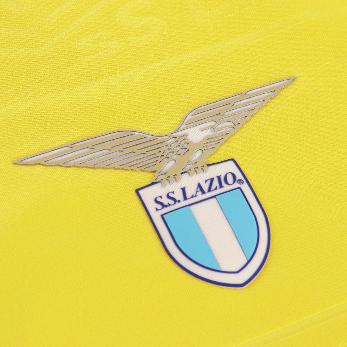 Lazio football club crest with a silver winged emblem on a bright yellow jersey background.