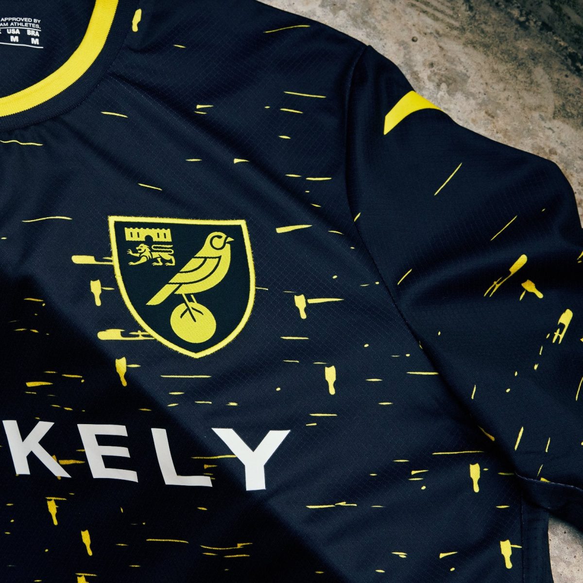 Close-up of a navy sports jersey with yellow speckle pattern and a yellow crest featuring a bird on a ball inside a shield at the chest area.