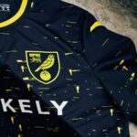 Close-up of a navy sports jersey with yellow speckle pattern and a yellow crest featuring a bird on a ball inside a shield at the chest area.