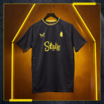 Dark athletic jersey on a hanger with yellow sponsor logo 'Stake.com' and Castore emblem; geometric pattern and neon yellow backdrop.