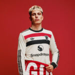 Person modeling a white jersey with red-black horizontal bands and Snapdragon logo, standing against a solid red background.