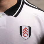 O273mF6UigPY8D4 | UAE SPORT STORE Close-up of a white polo shirt collar and chest, displaying a shield-shaped club logo with red initials on a white background and black border.