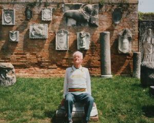 Older man in a Venezia sweatshirt sits on a stone block in a grassy yard, a brick wall with decorative stone reliefs behind him.