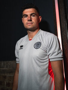 Portrait of a man in a white football jersey leaning against a wall at night, red light illuminating his side.