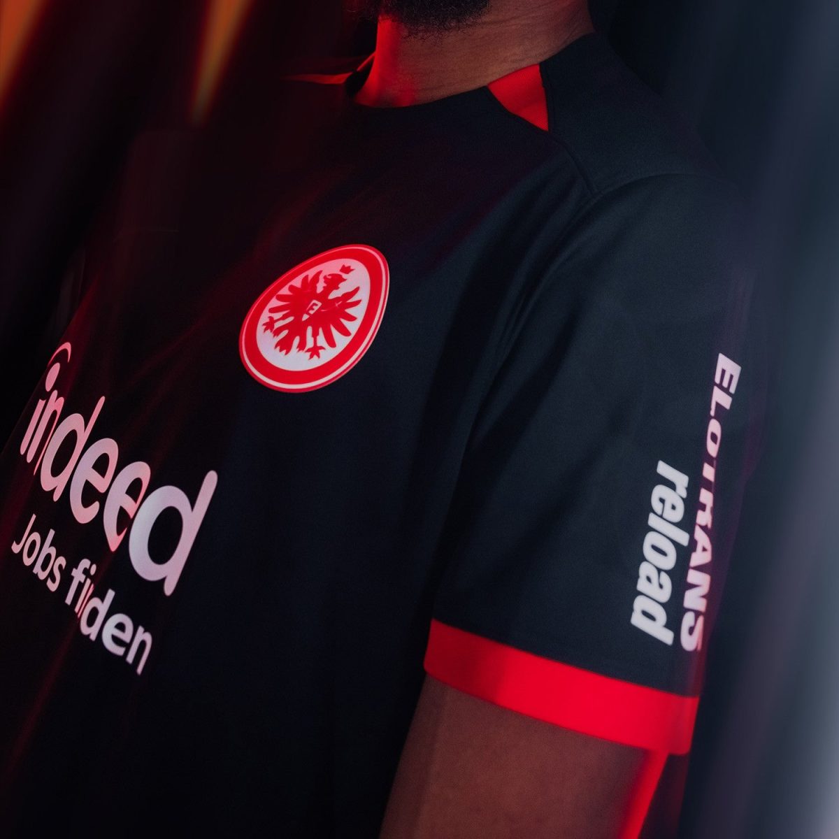 Close-up of a person wearing a black shirt with red trim; a circular red-and-white badge with an eagle on the chest, and white text ('indeed') partially visible on the front.