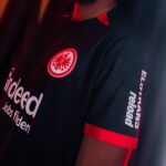 ObaiISkzyLMJp6V | UAE SPORT STORE Close-up of a person wearing a black shirt with red trim; a circular red-and-white badge with an eagle on the chest, and white text ('indeed') partially visible on the front.