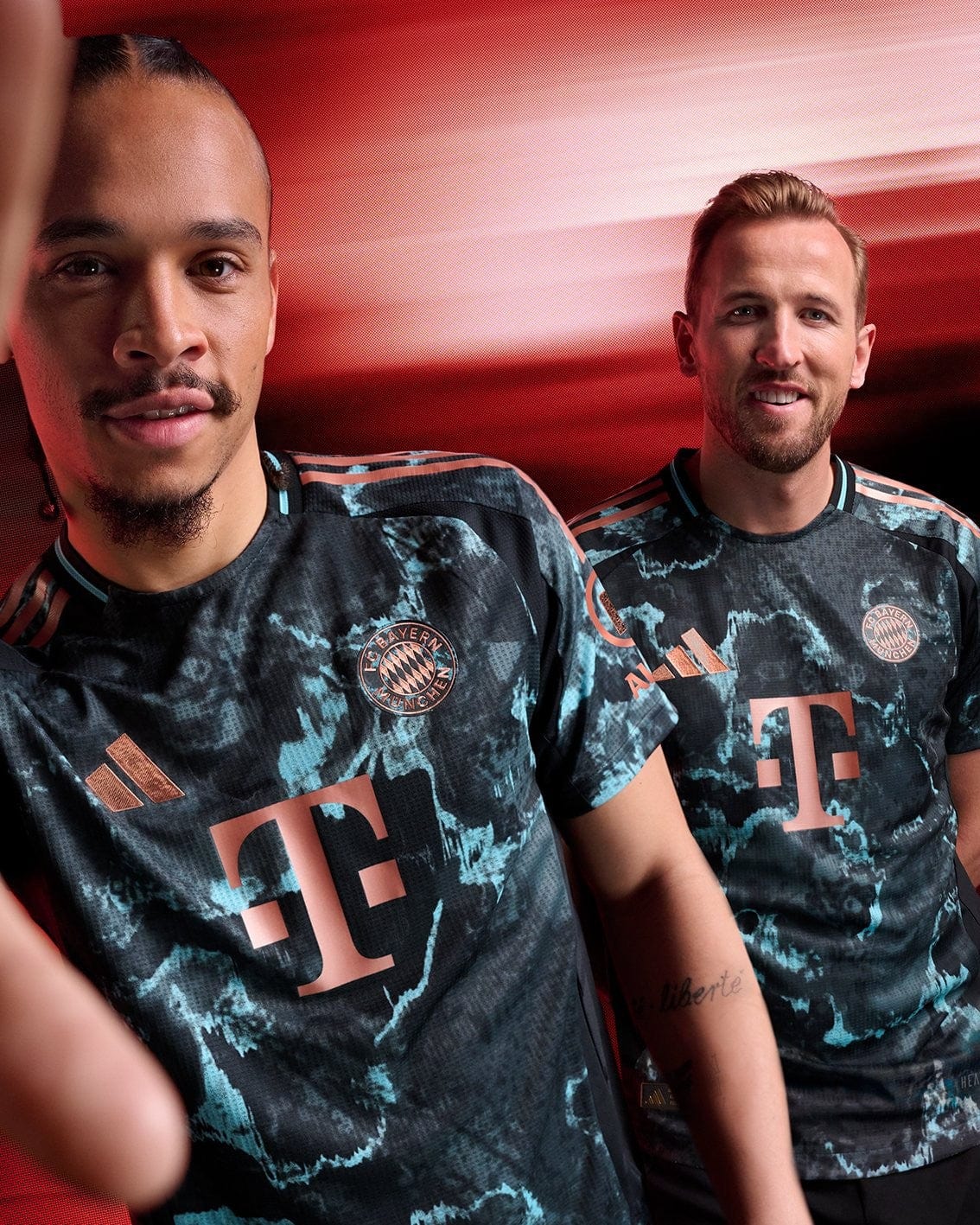 OxWg4eciwtmDKB7 | UAE SPORT STORE Two men in Bayern Munich training jerseys pose together against a red background, smiling at the camera with teal and copper accents on their shirts.