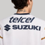 Back of a person wearing a white shirt with navy 'telcel' and 'SUZUKI' logos and Suzuki emblem; tattoo on the neck and a hoop earring visible.