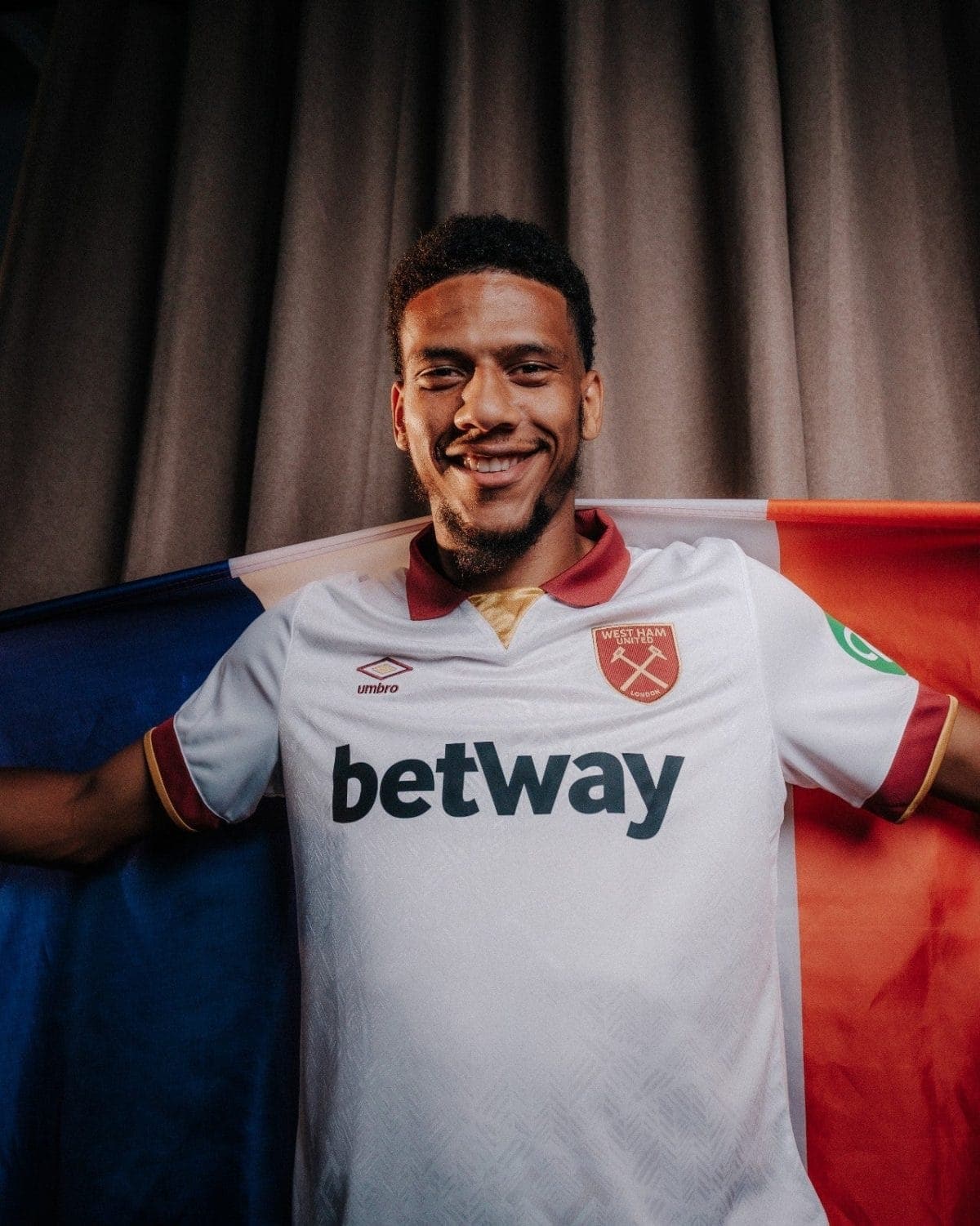 Smiling man in a West Ham United jersey posing with a blue-red flag behind him.