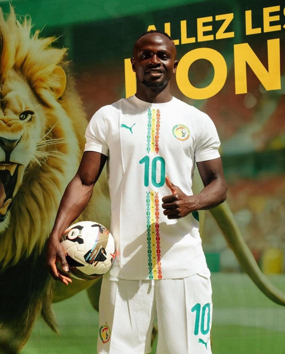 Soccer player in white Senegal kit with number 10, holding a soccer ball and giving a thumbs up, stands in front of a lion-themed backdrop.