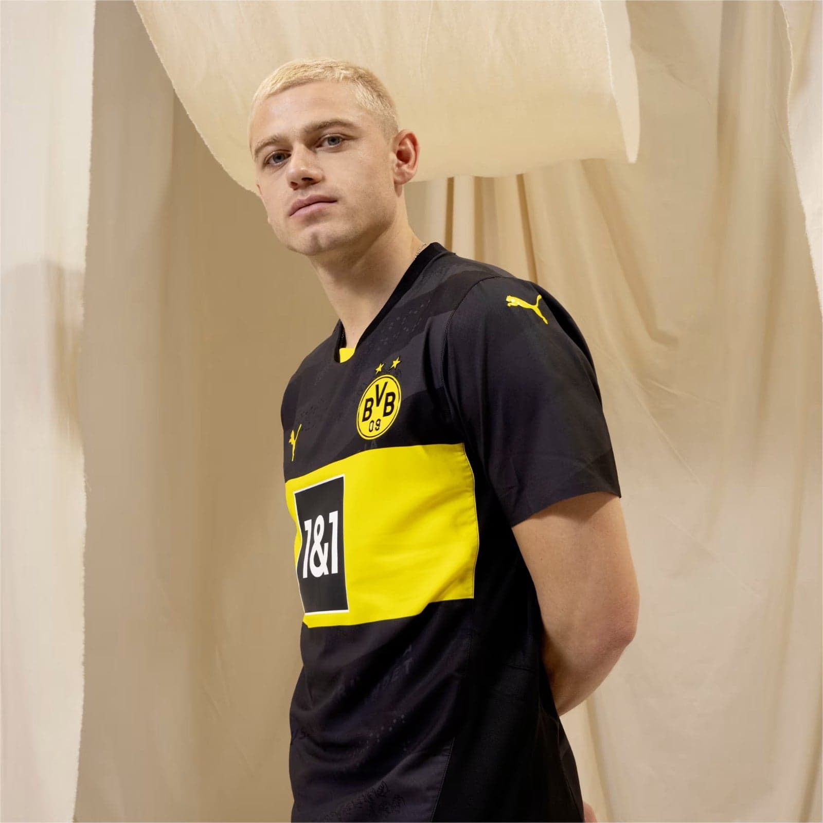 PMM4TXxkvee4q9w | UAE SPORT STORE Young man with dyed blond hair wearing a Borussia Dortmund black and yellow jersey, standing against beige curtains.