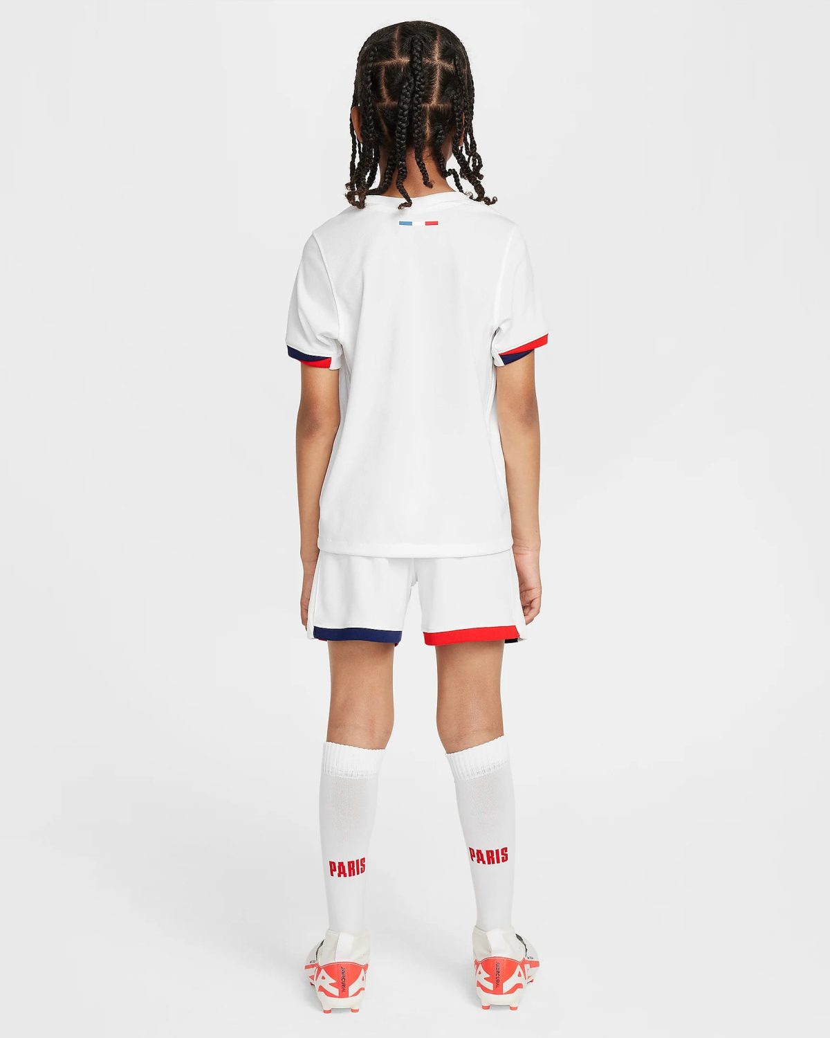 Back view of a young athlete in a white soccer jersey and shorts with red/blue trim, red 'PARIS' text on white socks, standing against a light background.