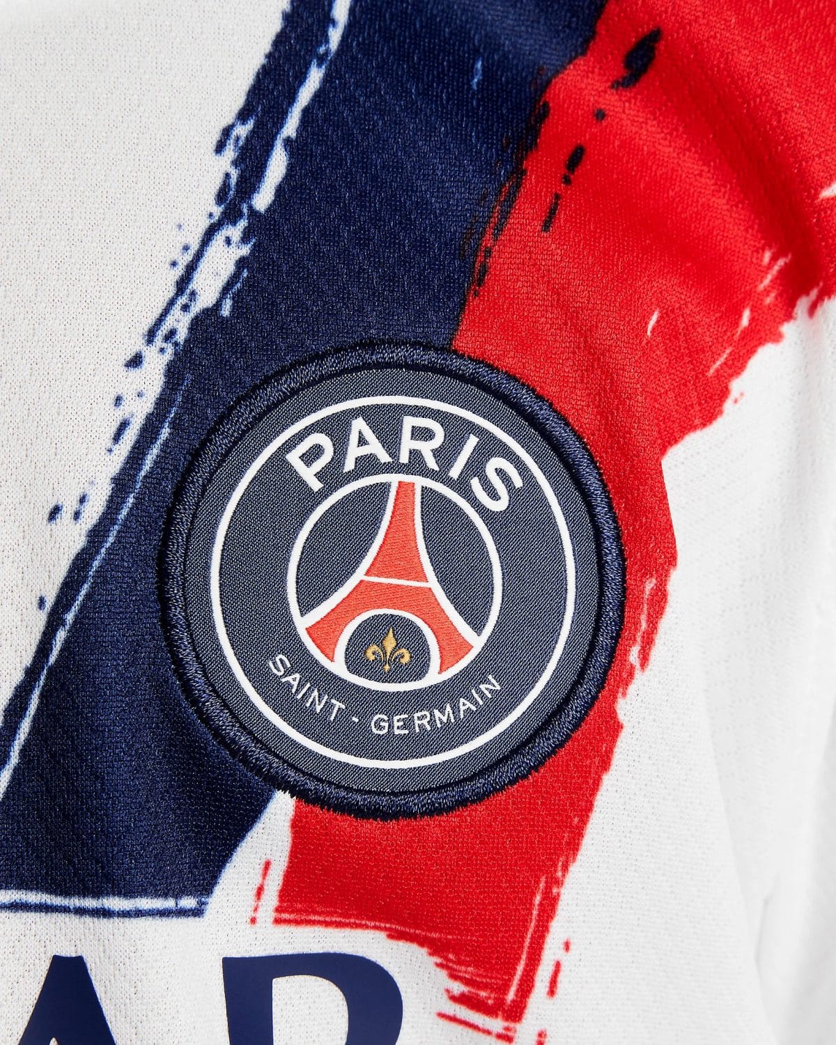 Close-up of a Paris Saint‑Germain patch on fabric, with dark blue and red paint-like streaks behind it.