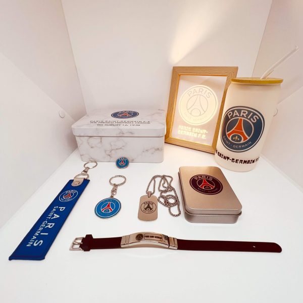 PSG-branded collection on a white shelf: box, framed logo, tumbler, keychains, lanyard, tin mint, and jewelry pieces.