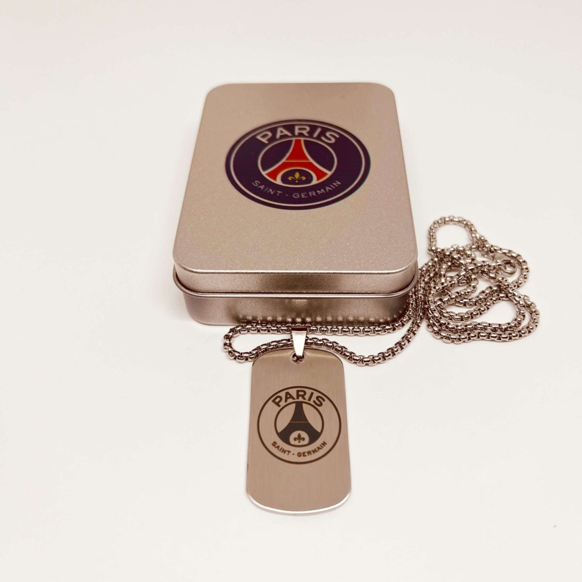 Rose-gold pendant with a Paris Saint-Germain logo on a linked chain resting on a metallic jewelry tin.