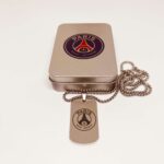 Paris-Saint-Germain-Dog-Tag-Titanium-Steel-Necklace-scaled-1 | UAE SPORT STORE Rose-gold pendant with a Paris Saint-Germain logo on a linked chain resting on a metallic jewelry tin.