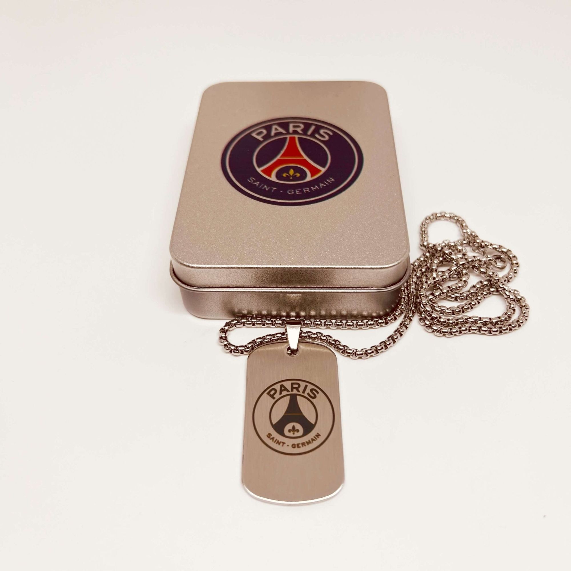 Paris-Saint-Germain-Dog-Tag-Titanium-Steel-Necklace-scaled-1 | UAE SPORT STORE Rose-gold pendant with a Paris Saint-Germain logo on a linked chain resting on a metallic jewelry tin.