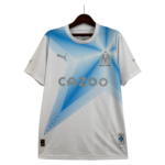 White soccer jersey on a hanger with blue chevron gradient, featuring OM crest and 'DROIT AU BUT' slogan on the chest, and 'CAZOO' sponsor text across front