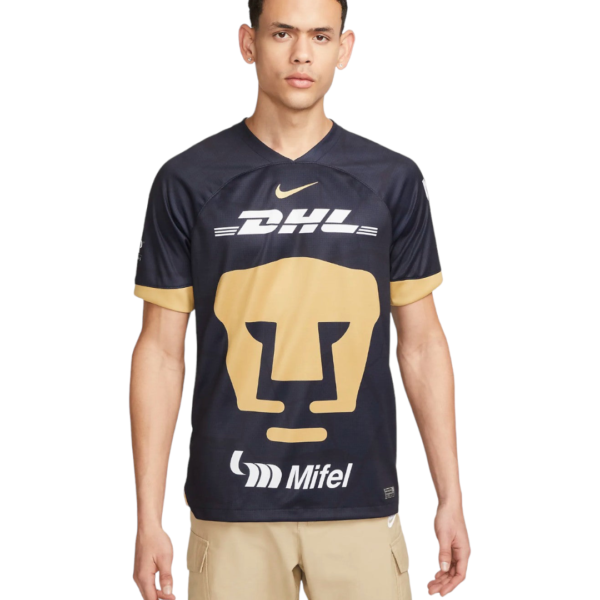 Young man wearing a dark sports jersey with a large gold face logo and DHL and Mifel logos, beige shorts, neutral pose