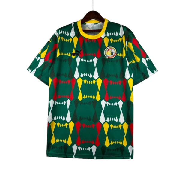 Green geometric-pattern soccer jersey on a hanger with red, yellow, and white diamond shapes, yellow collar, and a circular crest on the chest along with a small brand logo on the opposite side.